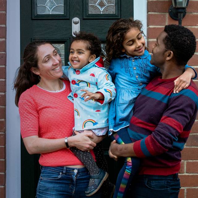 A white mother and Black father each hold one of their two daughters on the front doorstep of their home. The family is smiling at each other.