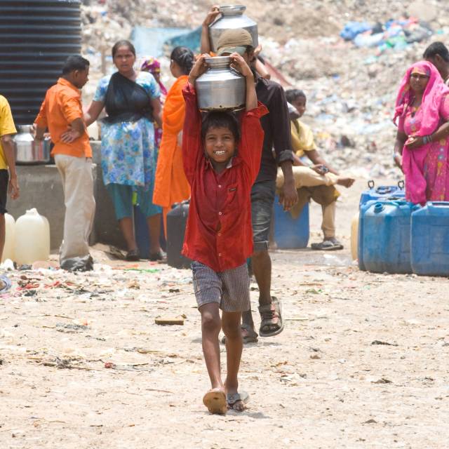 A young boy in India carries a metal water jug on his head. He is wearing a red button-up shirt, grey shorts and sandals.