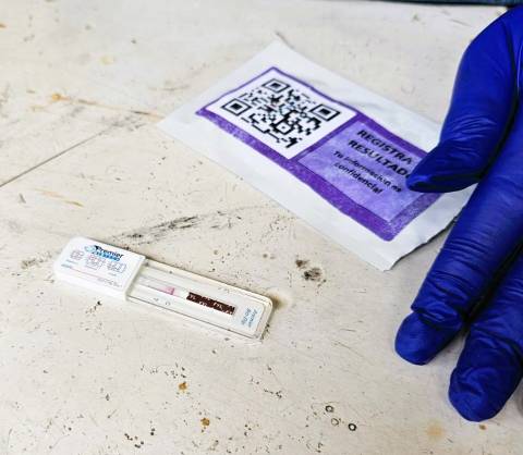 Blue gloved hand touches a fentanyl test strip wrapper with the test revealed underneat.