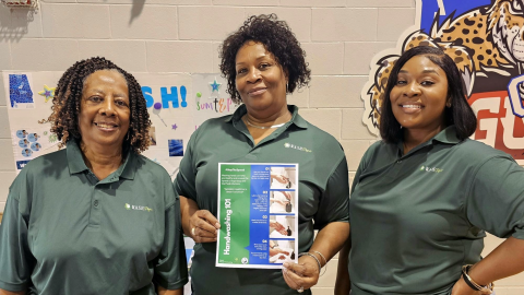 Three black women in green polos hold up a paper that says "handwashing 101"