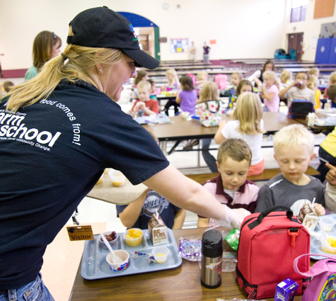 A person handing out food in a school lunchroom. Children are seated at lunchtables.