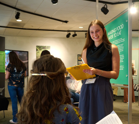 A smiling student writing on a clipboard and talking to a seated CDC scientist