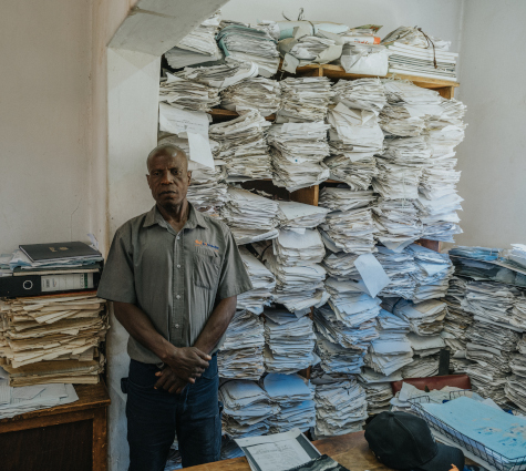 A man standing in font of a shelving unit that is overflowing with paper.