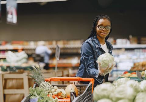 A young Black woman wearing glasses examines a head of lettuce in a grocery store