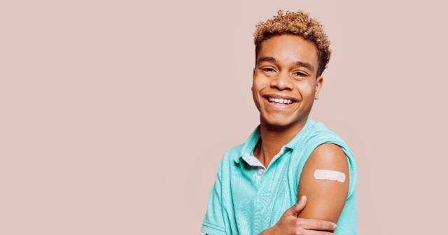 A young man smiles while showing a bandaid on his arm. 