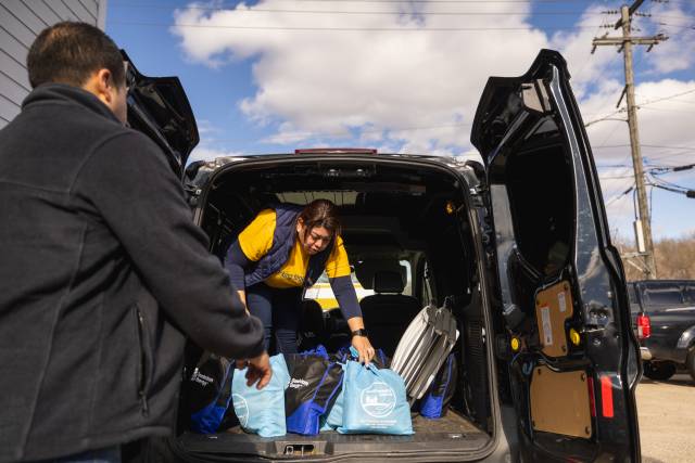 A woman in a yellow shirt and blue vest lifts bags out of the trunk of a van.