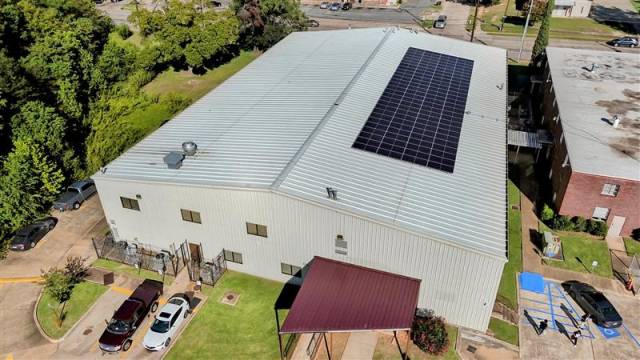 Aerial view of a large rectangular warehouse with solar panels on the roof, surrounded by greenery and parking spaces with several cars parked.