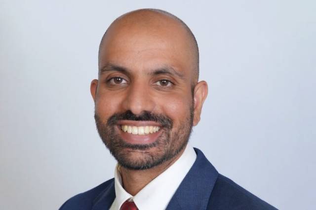 A headshot of a bald man with a beard and a blue suit and red tie.