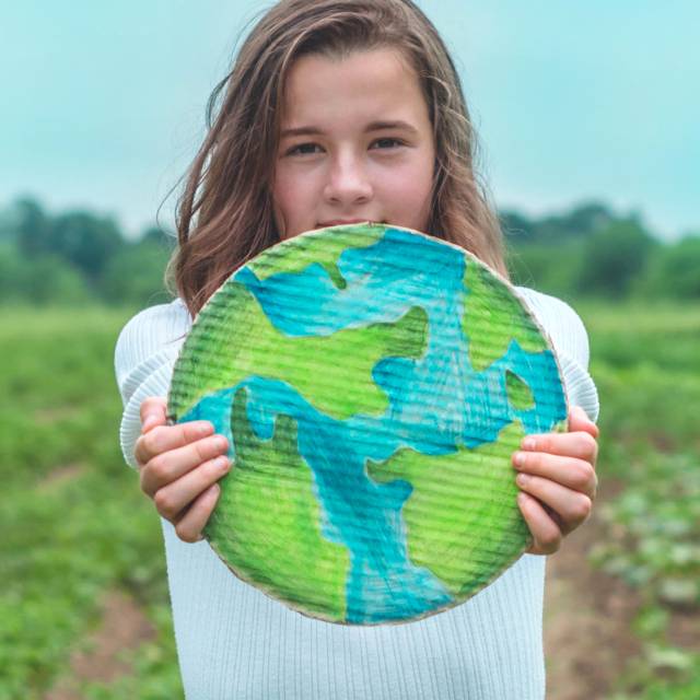 A young white girl with long brown hair stands in a field, holding out a handmade cardboard representation of the Earth.