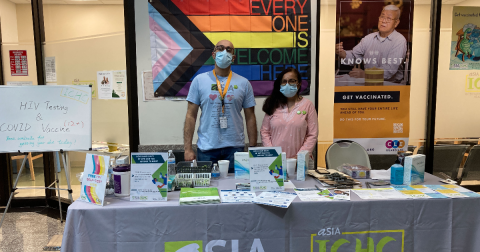 Two people stand at a table offering HIV self-testing materials. A pride flag hangs behind them.