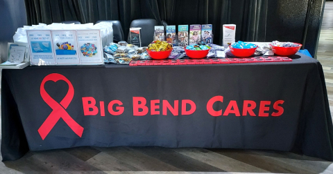 A table with HIV self-testing materials on it covered in a tablecloth with the words "Big Bend Cares" and HIV awareness ribbon