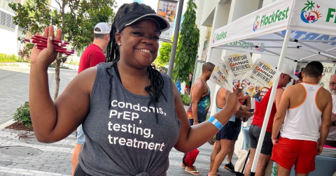 A woman in a gray shit that says "condoms, PrEP, testing, treatment" holds up pamphlets and pens.