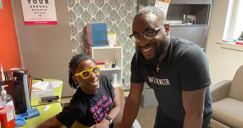 Two people in black t-shirts smile while sitting in an office.