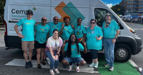 A group of people wearing teal shirts that say "CEMPA" stand in front of a white van and smile.