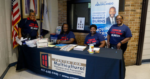 Four women in navy shirts that say "Caribbean Health Summit volunteer" smile while standing behind a table with a tablecloth that says "Center for Multicultural Wellness and Prevention"