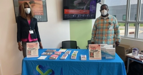 Two people in masks stand behind a table with blue tablecloth covered in HIV self testing educational materials and kits.