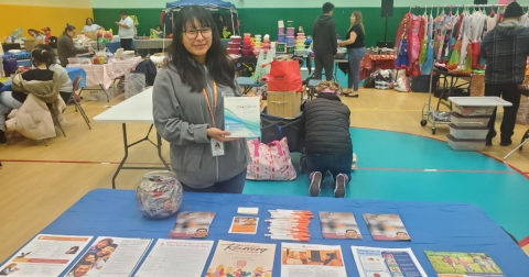 A woman in glaces holds up an HIV self-testing kit while standing behind a table with different reading materials on it.