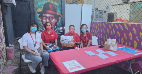 Four people wearing masks hold up HIV self-testing materials while standing behind a red table and in front of a mural
