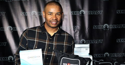 A man wearing a black button down shirt poses with HIV self-testing kits in front of a banner that says "sex ready."