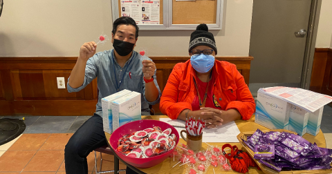 Two people in masks sit at a table filled with HIV self-testing kits, condoms, lollipops, and other swag. A man holds up heart shaped lollipops.