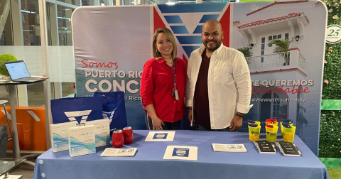 A man in a white button down and a woman in a red button down pose behind a table with HIV self-testing kits and flyers on it.
