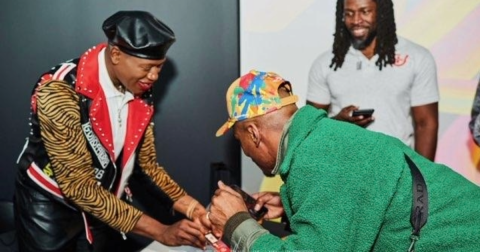 Three people crowd around a picture. One man wears a green sweatshirt and colorful hat. Another person wears a leopard print leather jacket and leather cap.