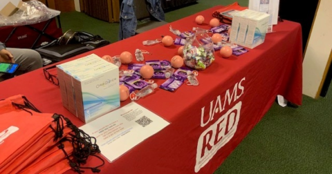 A table with a red table cloth that says UAMS has HIV self-testing kits, condoms, hand sanitizer and stress balls on it.
