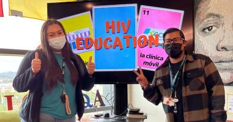 Two people in masks give a thumbs up and peace sign while standing in front of a monitor that says "HIV Education"