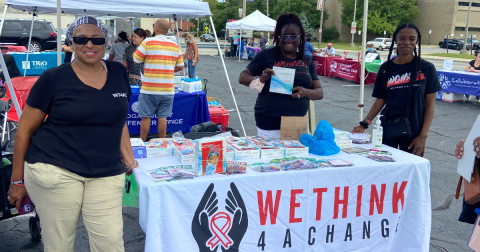 Three women in black shirts stand in front of a table with HIV self-testing kits on it. The tablecloth says "We Think 4 a Change"