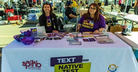 Two people wearing shirts that say "Big Auntie Energy" sit behind a table with HIV Self-testing materials placed on it.