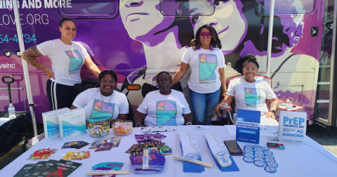 5 women in white tshirts with the Sister Love logo on them pose behind a table filled with HIV self-testing pamphlets and materials