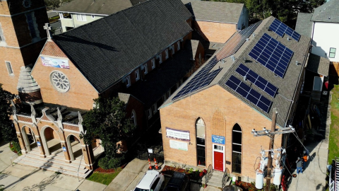Aerial view of a church featuring solar panels installed on its roof, surrounded by greenery and other buildings.