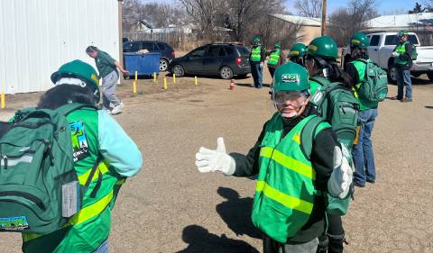 A young woman in a green emergency vest waving at the camera.