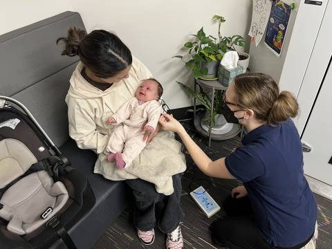 A hearing screener interacts with a newborn while the newborn's mother holds her.